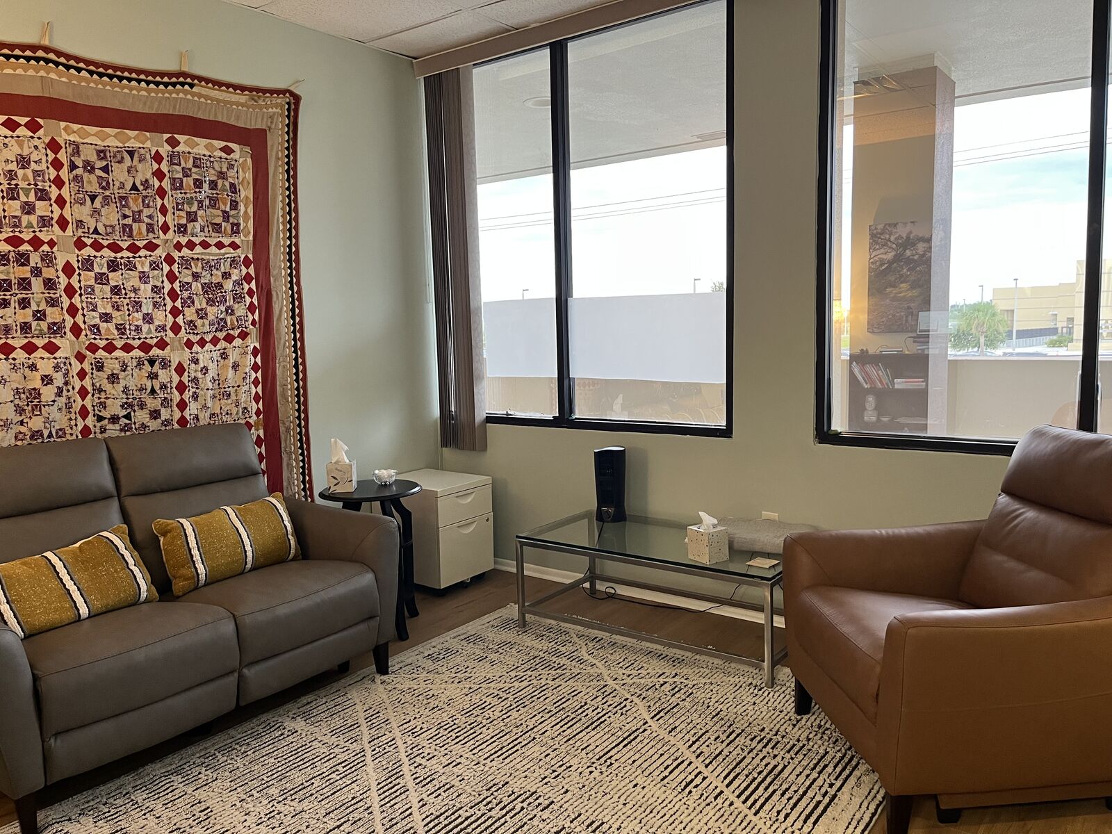 Therapy office with a gray sofa, leather armchair, textile wall hanging, and large windows
